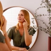 Frau vor dem Spiegel, die auf eine entzündete Stelle in ihrem Gesicht zeigt