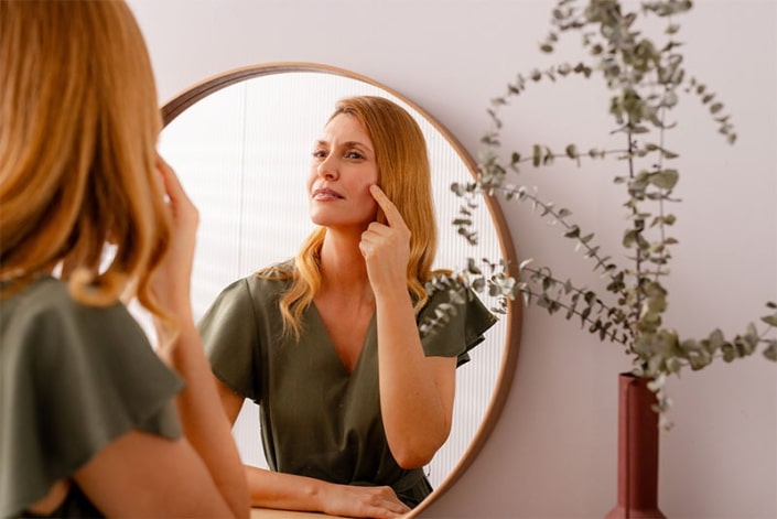 Frau vor dem Spiegel, die auf eine entzündete Stelle in ihrem Gesicht zeigt